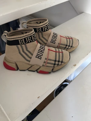 Burberry beige sneakers - Säljer ett par beige sneakers från Burberry med klassiskt rutmönster, svarta och röda detaljer samt bred resår med Burberry-logga över foten. Skorna har en chunky sula med röda inslag och stickad ovandel som ger en strump-liknande passform. Utan box