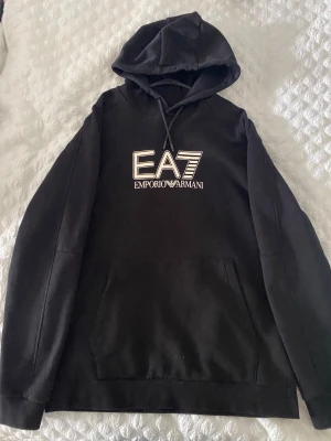 Svart EA7 Emporio Armani hoodie - Snygg svart hoodie från EA7 Emporio Armani med stor logga på bröstet. Klassisk känguruficka framtill, huva med dragsko och långa ärmar. Tillverkad i mjukt bomullsmaterial som känns skönt mot huden. Perfekt för en avslappnad och trendig look.
