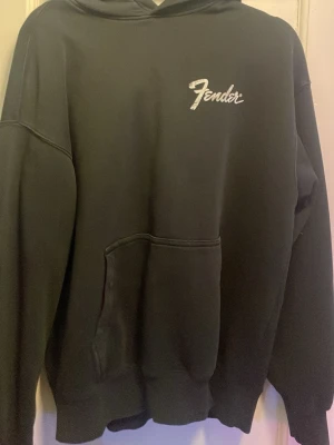 Svart Fender hoodie med tryck - Säljer en svart hoodie från Fender med vit logga på bröstet och stort tryck av en Deluxe Reverb-amp på ryggen. Hoodien har huva och känguruficka framtill. Perfekt för dig som gillar musik och vill ha en skön, avslappnad stil.