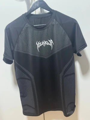 YOUNGLA T-shirt - Säljer en svart teknisk t-shirt från YOUNGLA med sportig design och tryckt logga på bröstet. T-shirten har korta ärmar och är tillverkad i ett lätt och ventilerande syntetmaterial, perfekt för träning eller en aktiv vardag.