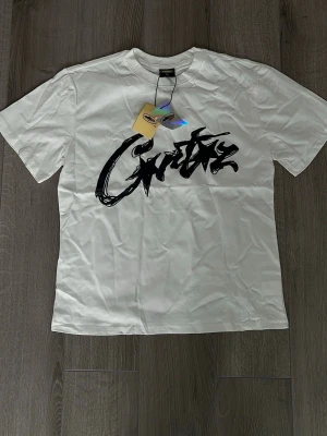 Corteiz vit tshirt  - Säljer en vit t-shirt från corteiz med ett stort svart graffiti-inspirerat tryck på bröstet. Klassisk rund hals och korta ärmar. Tillverkad i mjuk bomull för skön känsla. Perfekt för dig som gillar streetwear och vill sticka ut.