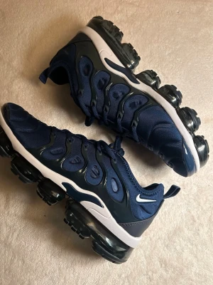 Nike Air Vapormax Plus blå sneakers - Nike Air Vapormax Plus sneakers i mörkblå mesh och syntet med svarta och vita detaljer. Skorna har en unik, synlig Air-sula i svart och en vågig design på ovandelen. Perfekta för dig som gillar sportig och modern stil. Swoosh-logga på sidan och Airmax-tryck under sulan.