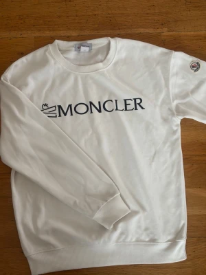 Vit sweatshirt från Moncler - Snygg vit sweatshirt från Moncler med svart logotyptryck på bröstet och Moncler-märke på ärmen. Tröjan har rund halsringning, långa ärmar och ribbade muddar. Perfekt för en clean och stilren look.