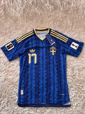 Blå Sverige fotbollströja Adidas - Säljer en blå Sverige fotbollströja från Adidas med gula detaljer och nummer 17 på bröstet. Tröjan har officiellt Sverige-emblem, gula ränder på axlarna och patchar på ärmarna. Tillverkad i lätt och ventilerande material, perfekt för match eller träning.