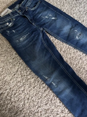 Blå slitna jeans från dondup - Säljer ett par blå jeans från Dr. Denim med snygga slitningar och tvättade detaljer. Modellen har smal passform och klassiska fem fickor. Jeansen är gjorda i bomull med lite stretch för extra komfort.