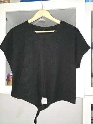 Svart ribbad topp med knyt - Svart ribbad topp med korta ärmar och rund halsringning. Toppen har en snygg knytning framtill som ger en avslappnad vibe. Perfekt att matcha med höga jeans eller kjol för en trendig look.