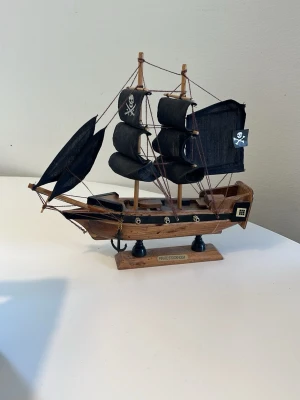 Pirat skepp modell - Säljer en detaljerad modell av ett piratskepp i trä med svarta segel och dödskallemotiv. Skeppet har små kanoner, ankare och repdetaljer. Perfekt som dekoration eller samlarobjekt.