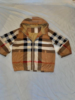 Burberry windbreaker  - Snygg beige vindjacka från Burberry med klassiskt rutmönster i svart, vitt och rött. Jackan har huva, dragkedja framtill och elastiska muddar vid ärmar och midja. Perfekt för dig som gillar streetstyle och vill sticka ut med ett ikoniskt mönster.