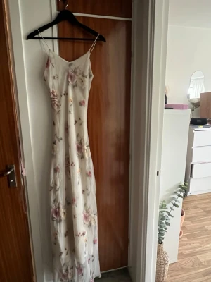 Lång blommig sommarklänning - Säljer en romantisk långklänning med tunna axelband och vackert blommönster i pastellfärger på vit botten. Klänningen har en smäcker och skön passform. Materialet känns lätt och svalt, vilket gör den superbekväm att bära.