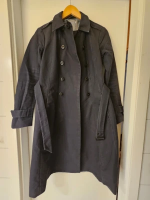 G-Star RAW trenchcoat med skärp – stl S - Säljer en klassisk och stilren trenchcoat från G-Star RAW i mörkblå färg. Jackan har en tidlös design med midjebälte och passar perfekt för vår, höst och mildare vinterdagar.  Bra begagnat skick. Jackan är använd och har normalt slitage samt vissa veck efter användning, men inga hål, revor eller andra skador. Alla delar är hela och fungerar som de ska.