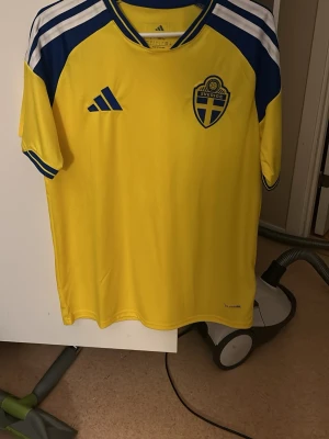 Svensk landslagströja Adidas gul/blå - Gul och blå fotbollströja från Adidas med Sveriges landslagsemblem på bröstet och klassiska Adidas-ränder på axlarna. Tröjan har korta ärmar, rund hals och är tillverkad i ett lätt, ventilerande material. Perfekt för match eller träning.