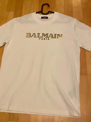 Vit Balmain Paris t-shirt i bomull - Snygg vit t-shirt från Balmain Paris med guldfärgat tryck på bröstet. Klassisk rund halsringning och korta ärmar. Tillverkad i mjuk bomull för en skön känsla och stilren look.