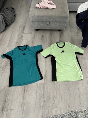 Adidas domartröjor turkos & ljusgrön - Två domartröjor från Adidas, en i turkos och en i ljusgrönt med svarta sidopaneler och detaljer. Båda är storlek M och har två stora bröstfickor. Tillverkade i lätt, syntetiskt material med AEROREADY-teknologi för bra ventilation. Perfekta för fotbollsdomare.