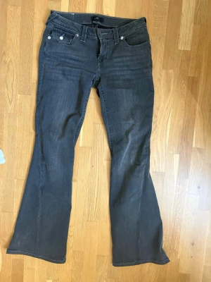 Gråa bootcut jeans från True Religion - Säljer ett par svarta bootcut jeans från True Religion med klassiska fickor bak och snygga silverknappar❤️. Jeansen har låg midja och coola sömdetaljer. Stl 27. Använda fåtal gånger och nypris är 1399kr🥰