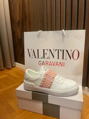 Valentino Garavani rockstud vita sneakers - Stilrena vita sneakers från Valentino Garavani med en bred ljusrosa rem över vristen dekorerad med ikoniska nitar. Skorna har en klassisk låg siluett, rund tå och platt vit sula. Perfekta för dig som vill ha en exklusiv touch på din outfit. Skriv gärna om det är något du undrar över