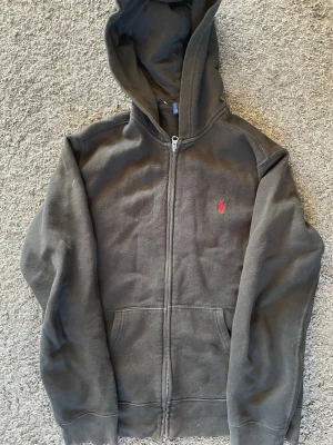 Svart hoodie från Polo Ralph Lauren - Klassisk svart hoodie från Polo Ralph Lauren med dragkedja framtill och huva. Ikoniska röda loggan broderad på bröstet och två fickor fram. Perfekt för en chill och stilren look.