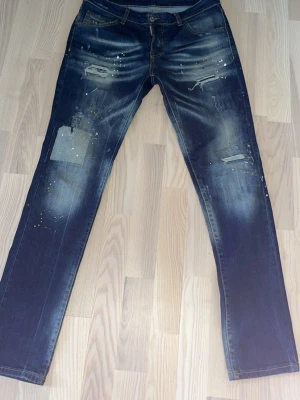 Dsquared2 slitna blå jeans herr - Säljer ett par blå jeans från Dsquared2 med slitningar, patchade detaljer och färgstänk. Jeansen har rak passform och klassisk femficksdesign. Snygga kontrastsömmar och låg midja ger en cool streetwear-look.