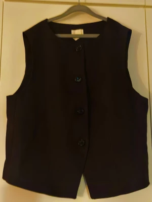 Mörkbrun väst med knappar från H&M - Stilren svart väst med fyra stora knappar framtill. Västen har en rak passform och är ärmlös med rundad halsringning. Perfekt att styla över en skjorta eller t-shirt för en clean look.