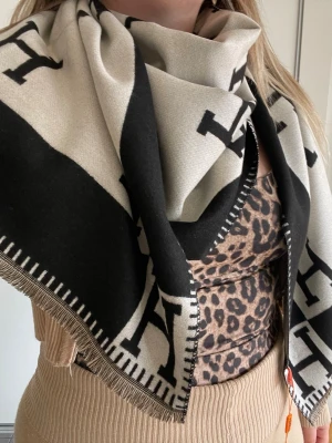 Beige och svart halsduk från H - Stilren halsduk från HERMS i beige och svart med stora bokstäver och dekorativa fransar längs kanten. Tjockt och mjukt material som känns lyxigt och passar perfekt för att lyfta din outfit. Fyrkantig form och klassisk design.