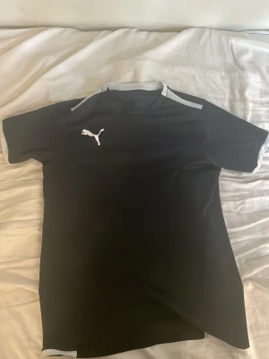 Svart Puma t-shirt med vita detaljer - Snygg svart t-shirt från Puma med vita detaljer på ärmslut och axlar samt Puma-logga på bröstet. Tillverkad i ett mjukt och lätt material som passar perfekt till träning eller chill. Klassisk passform och rund hals.