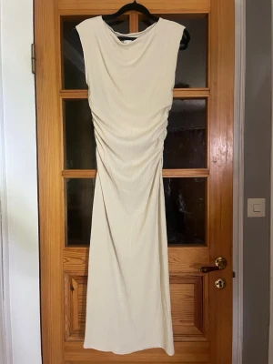 Lång beige klänning med rynk - Säljer en elegant långklänning i beige med rynkade detaljer längs midjan. Klänningen är ärmlös och har en mjuk, följsam passform som framhäver siluetten. Perfekt för dig som gillar stilrena och tidlösa plagg.