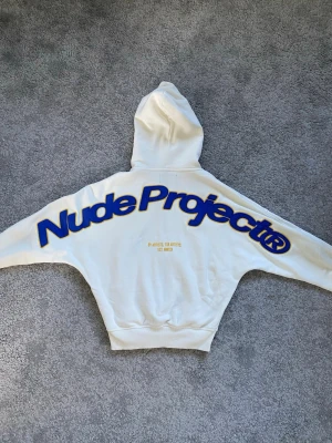 Vit hoodie från Nude Project - Säljer en vit hoodie från Nude Project med stor blå och gul logga på ryggen och text på bröstet. Hoodien har dragkedja, huva och ribbade muddar. Materialet är mjukt och bekvämt, perfekt för en avslappnad look. Priset kan diskuteras!