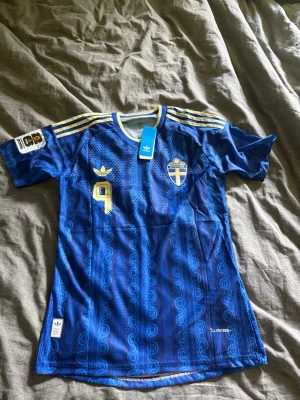 Blå Sverige fotbollströja Adidas - Säljer en blå Sverige fotbollströja med gult tryck, nummer 9 och namnet ISAK på ryggen. Tröjan har korta ärmar, guldfärgade detaljer och det svenska landslagets emblem på bröstet. Tröjan är aldrig använd och därför 10/10 i skick. Vid frågor hör av er! 