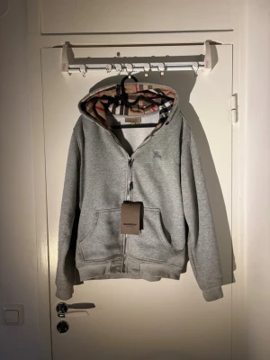 Grå zip hoodie Burberry - Snygg grå hoodie från Burberry med dragkedja och klassiskt rutigt foder i huvan. Hoodien har två stora fickor framtill och ribbade muddar vid ärmar och nederkant. Perfekt för en avslappnad och stilren look.