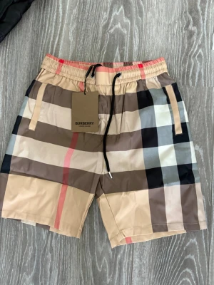 Beige rutiga shorts från Burberry - Säljer ett par klassiska beige shorts från Burberry med det ikoniska stora rutmönstret i svart, vitt, brunt och rött. Shortsen har elastisk midja med snörning och sidofickor. Perfekta för dig som vill ha en stilren och trendig look.