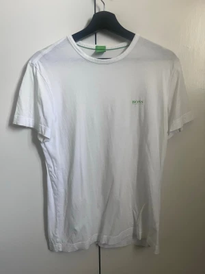 Vit t-shirt från Hugo Boss - Klassisk vit t-shirt från Hugo Boss med diskret logga i grönt på bröstet och större logga på ryggen vid axeln. T-shirten har rund halsringning och korta ärmar. Tillverkad i mjuk bomull för en skön känsla och enkel stil.