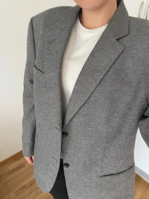 Grå oversized kavaj - Stilren grå kavaj i oversized modell med breda axlar och två knappar framtill. Kavajen har klassiska slag, två framfickor och en bröstficka. Materialet är vävt tyg som ger en elegant look. Hittar ingen storlek men passar bra på mig som är en S/M, köpt second hand.