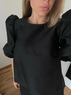 Svart blus med puffärm - Svart blus med puffiga trekvartsärmar och rund halsringning. Blusen har en rak passform och är tillverkad i ett slätt tyg som ger en stilren look. Perfekt för dig som gillar statement-ärmar och enkel design. Använd max 2 ggr