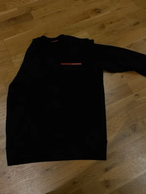 Prada tröja - Stilren svart Prada-tröja med exklusiv röd logodetalj som ger en perfekt balans mellan minimalistisk design och premiumkänsla. Bekväm passform, hög kvalitet och en look som lyfter varje outfit. En självklar favorit för dig som uppskattar diskret lyx och modern stil.  ✔ Elegant ✔ Bekväm ✔ Lätt att matcha ✔ Eftertraktad designerpiece