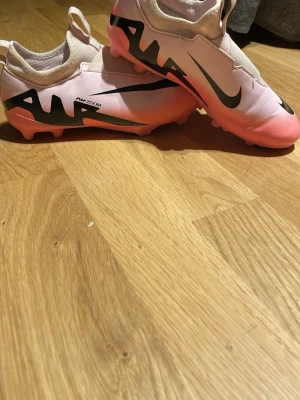 Nike Air Zoom rosa fotbollsskor - Säljer ett par Nike Air Zoom fotbollsskor i vitt och rosa med stora svarta detaljer och logga på sidan. Skorna har fasta dobbar och en modern, strömlinjeformad design. Ovandelen är i syntetmaterial och sulan är i rosa plast. Perfekta för dig som vill sticka ut på planen!