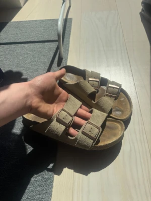 Beige Birkenstock Arizona sandaler storlek 40 - Säljer ett par klassiska Birkenstock Arizona sandaler i beige mocka med två justerbara remmar och metallspännen. Fotbädden är formad för extra komfort och sulan är i kork. Perfekta för en avslappnad och trendig stil.