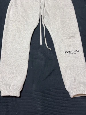 Gråa Essentials mjukisbyxor - Säljer ett par ljusgrå mjukisbyxor från Fear of God Essentials. Byxorna har resår i bensluten, dragsko i midjan och svart Essentials-tryck på benet. Tillverkade i mjukt bomullsmaterial som är perfekt för chill dagar.