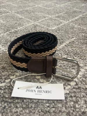 Randigt flätat bälte från John Henric - Snyggt flätat bälte från John Henric med randigt mönster i svart och beige. Bältet har detaljer i brunt skinn och ett klassiskt metallspänne. Perfekt accessoar för att lyfta din outfit.