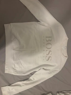 Vit collegetröja  från Hugo Boss - Säljer en stilren vit sweatshirt från Hugo Boss med stor logga framtill. Tröjan har rund halsringning och långa ärmar. Perfekt för dig som gillar klassisk och enkel design med en exklusiv touch.
