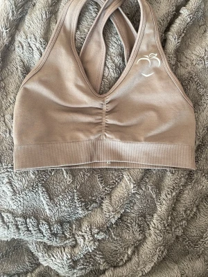 Beige sport-bh från Peachy - Säljer en beige sport-bh från Peachy med brottarrygg och diskret rynkning framtill. Bh:n har ett vitt persiko-tryck på ena sidan och bred resår under bysten för extra stöd. Tillverkad i stretchigt material som passar perfekt till träning.