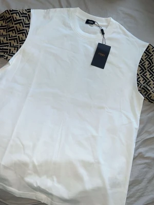 Vit Fendi t-shirt med logomönstrade ärmar - Snygg vit t-shirt från Fendi med korta ärmar i beige och svart logomönster. Klassisk rund halsringning och exklusiv känsla. Perfekt för dig som vill ha något trendigt och stilrent från ett lyxigt märke.