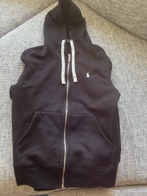 Svart  hoodie från Polo Ralph Lauren - Svart  hoodie från Polo Ralph Lauren med dragkedja framtill, vita snören i huvan och broderad logga på bröstet. Tillverkad i mjuk bomull och har två stora fickor framtill. Perfekt för en avslappnad streetwear-look.