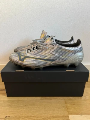 Mizuno silver fotbollsskor - Snygga fotbollsskor från Mizuno i silver med grå och gula detaljer. Skorna har en låg profil, snörning och mönstrad ovandel i syntetmaterial för bra bollkontroll. Perfekta för dig som vill sticka ut på planen med stilrena och lätta skor.