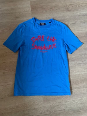 Blå t-shirt från scotch & soda - Blå t-shirt med trycket 'Surf the Soundwave' i rött på bröstet. Klassisk rund hals och korta ärmar. Tillverkad i mjuk bomull, perfekt för en avslappnad stil.