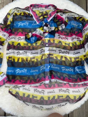 Färgglad transparent skjorta Made in the Shade (XS) - Säljer en transparent skjorta från Made in the Shade i storlek XS. Skjortan har färgglada ränder i rosa, blått, gult och svart med texttryck och knappar framtill. Materialet är tunt och lätt, perfekt för dig som vill sticka ut.
