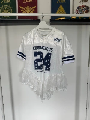 Vit jersey-tröja med volang  - Vit jersey-topp med blått tryck. Gjord i mesh och satinliknande polyester med sportiga ränder på ärmarna och volangdetalj nedtill, framhäver höfterna superfint!