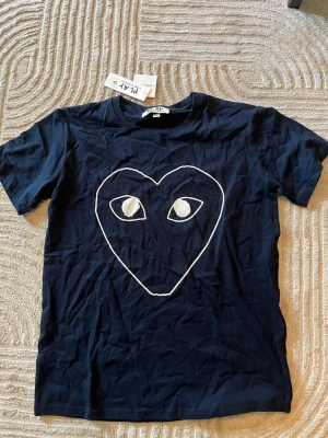 Comme des Garçons PLAY t-shirt - Snygg mörkblå t-shirt från Comme des Garçons PLAY med det ikoniska hjärtmotivet med ögon på bröstet. Klassisk passform och rund hals. Tillverkad i mjuk bomull, perfekt för en avslappnad och trendig look. Storlek XL men är väldigt liten i storlek så passar bättre som L och M. Oanvänd och Ny så pris går att diskutera!