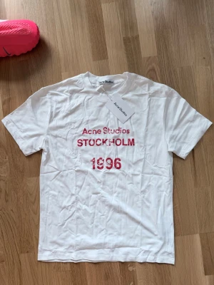 Vit Acne Studios t-shirt med tryck - Vit t-shirt från Acne Studios med rött tryck på bröstet där det står 'Acne Studios STOCKHOLM 1996'. Klassisk passform och rund hals. Tillverkad i mjuk bomull, perfekt för en clean och stilren look.