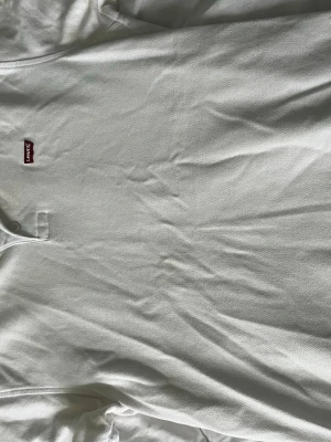 Vit t-shirt från Levi's med logga - Klassisk vit t-shirt från Levi's med den ikoniska röda loggan på bröstet. T-shirten har rund halsringning och korta ärmar. Tillverkad i mjuk bomull för en skön känsla och enkel stil.