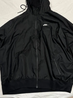 Svart vindjacka från Nike med huva - Snygg svart vindjacka från Nike med dragkedja framtill, två fickor med dragkedja och justerbar huva. Jackan har en stilren design med Nike-logga på bröstet och elastiska muddar vid ärmslut och nederkant. Perfekt för en sportig och avslappnad look.