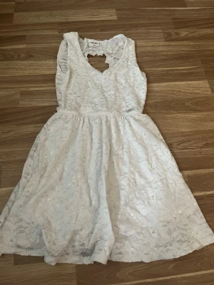 Vit spetsklänning med hjärtformad rygg - Söt vit klänning i spets med blommigt mönster och hjärtformad öppning i ryggen. Klänningen har v-ringning och är ärmlös, perfekt för sommarens alla tillfällen. Midjan är markerad och kjolen har en lätt utsvängd form.